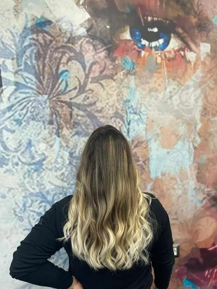 Capelli lunghi mossi, castani alla radice e biondi sulle punte, di spalle davanti a un grande volto di donna sulla parete